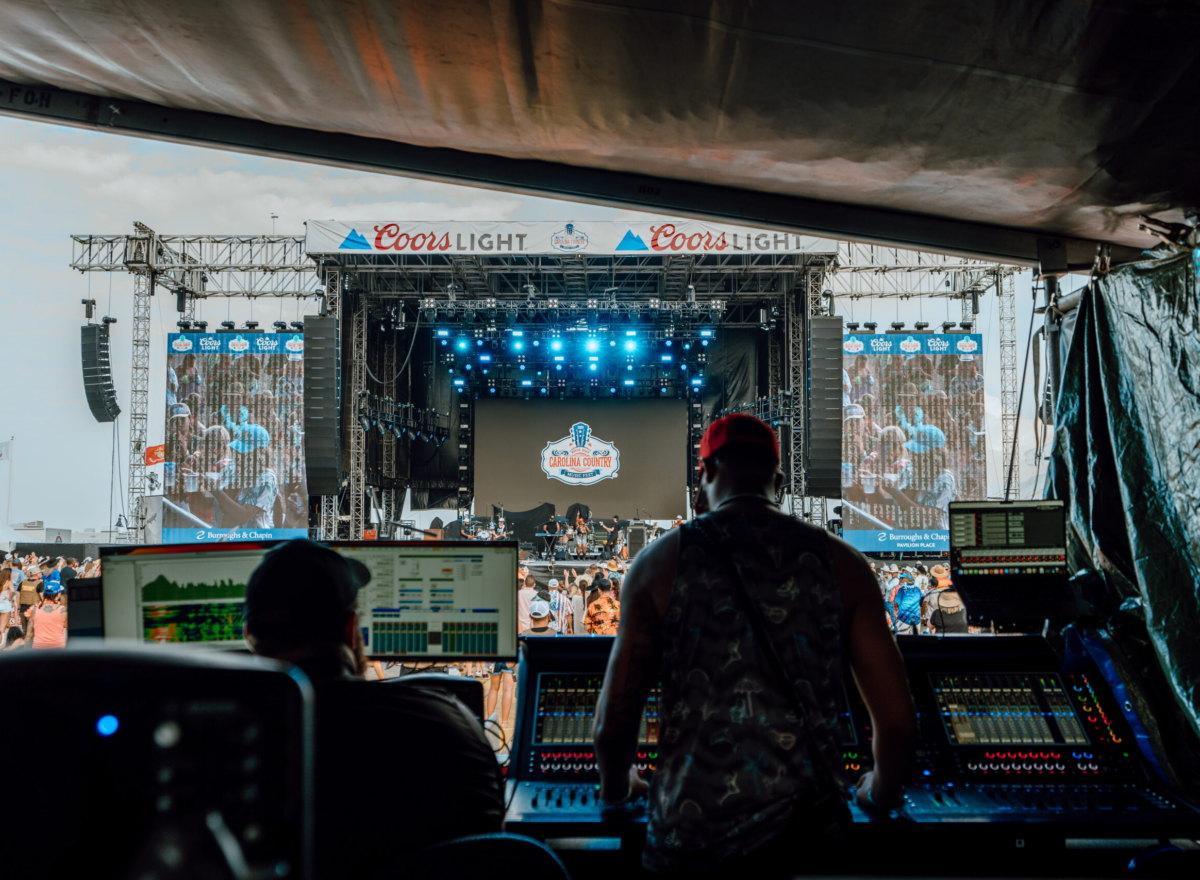 Front of House equipment and technicians with outdoor stage and crowd in the background.